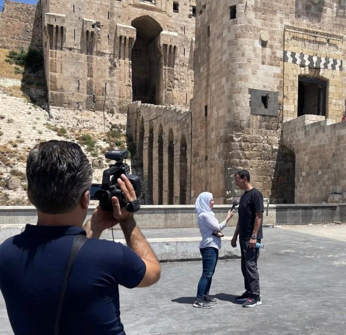 A person taking a picture of two people in front of a stone building

Description automatically generated