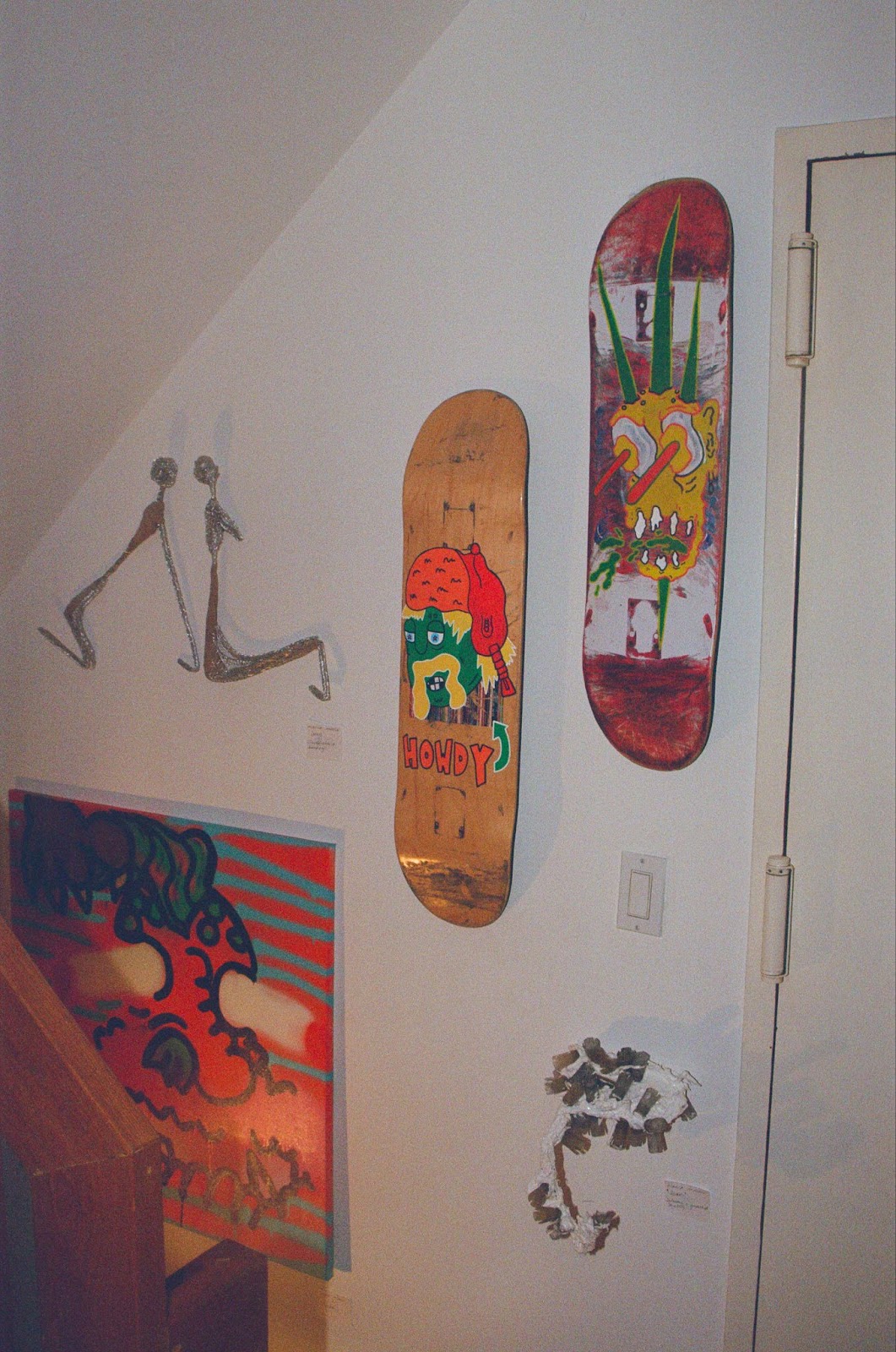 Five pieces of art hanging on a white wall: two painted skateboard decks, one chicken-wire sculpture, one plaster and bottle cap sculpture, and an orange and blue painting.