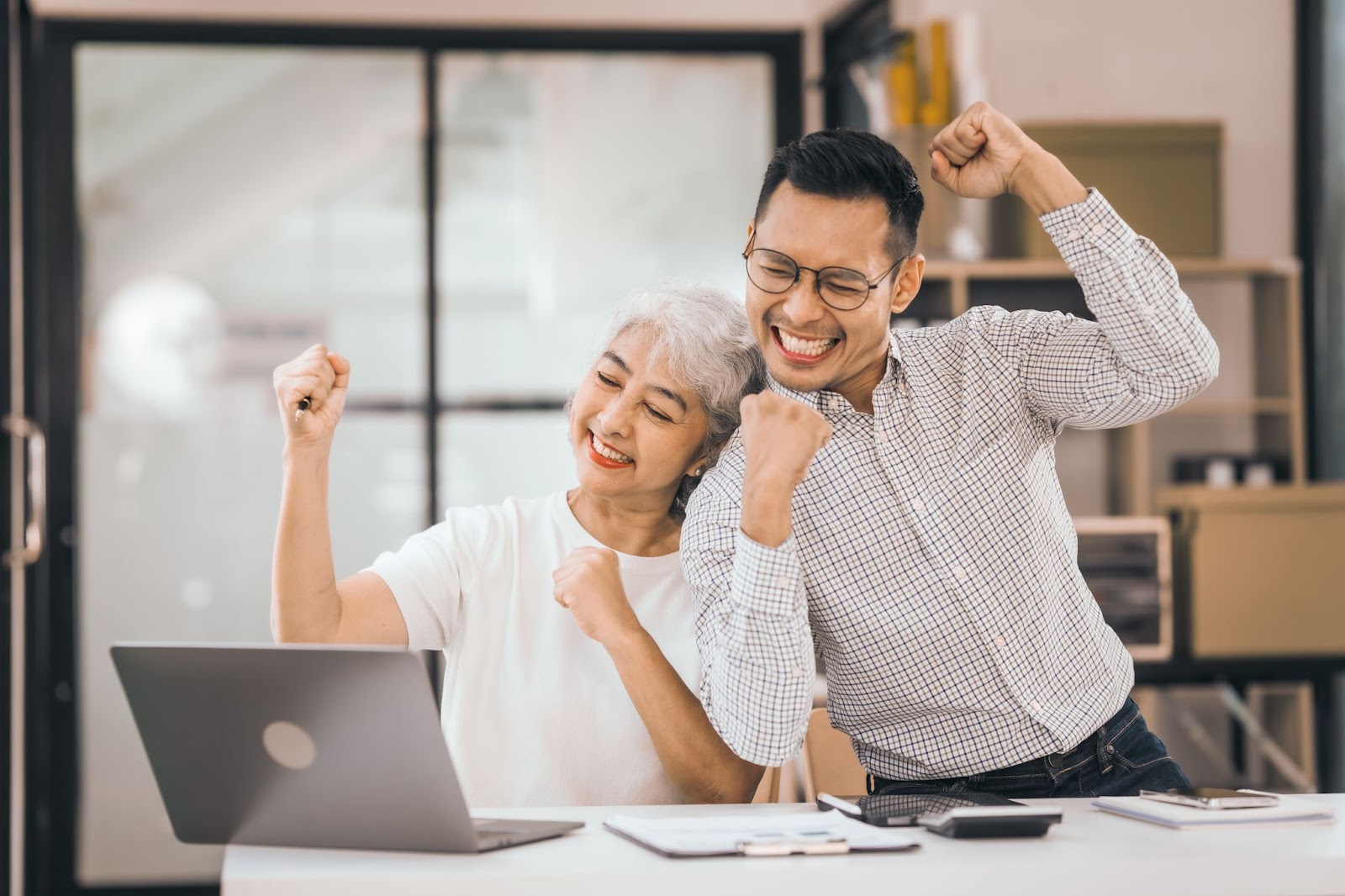 A mature woman and an Asian man joyfully review data on a laptop, appearing thrilled by the successful outcomes of a business strategy provided by a consultant.