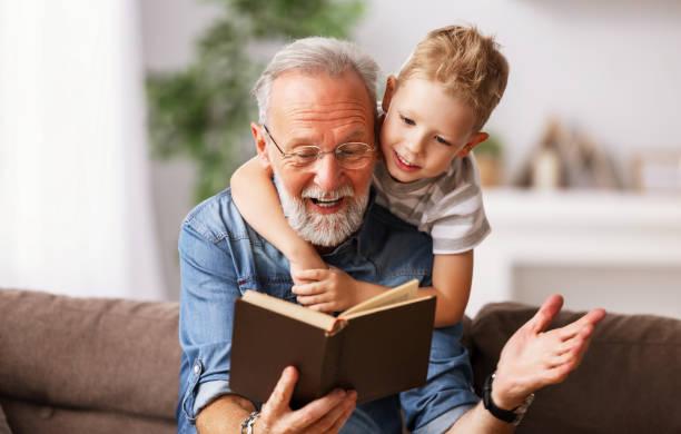 . Cheerful grandfather and grandson reading book together Happy family elderly  man and little boy smiling r while sitting on couch and reading fascinating fairy tale together at home boys growing up with grandfather stock pictures, royalty-free photos & images