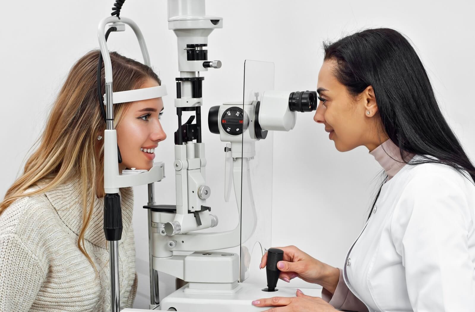 A young person with long blonde hair is receiving an eye exam from an optometrist, who is using a specialized machine to examine their eyes.