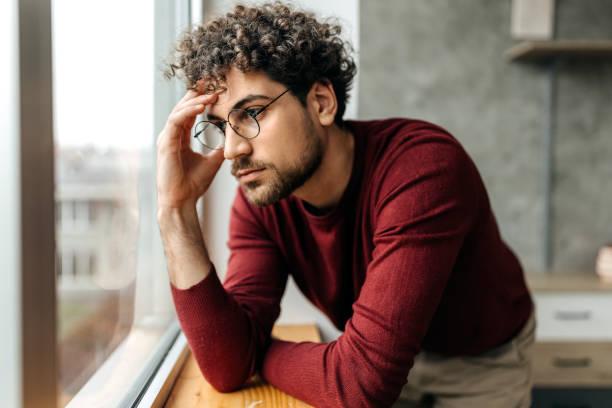 portrait of depressed man contemplating his thoughts while standing next to the window in the apartment - man in stress stock pictures, royalty-free photos & images