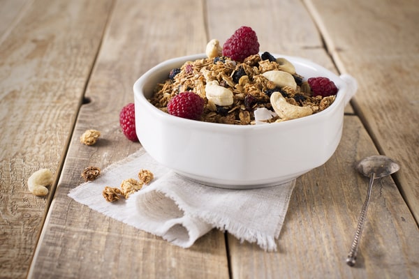 A bowl of cereals with raspberries, almond flakes, and coconut flakes placed on a wooden surface