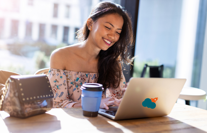 A smiling woman works on her laptop at a sunny cafe table, possibly researching the real estate market.