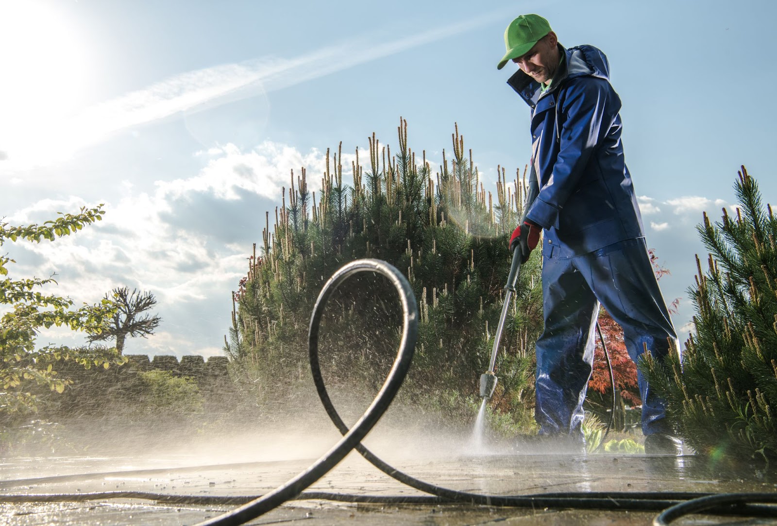 A gardener in blue protective gear and a green cap washing the asphalt pavement with a pressure washer.