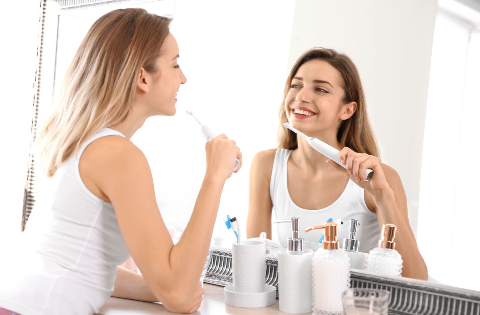 A woman looking into a bathroom mirror at herself while she is brushing her teeth with an electric toothbrush