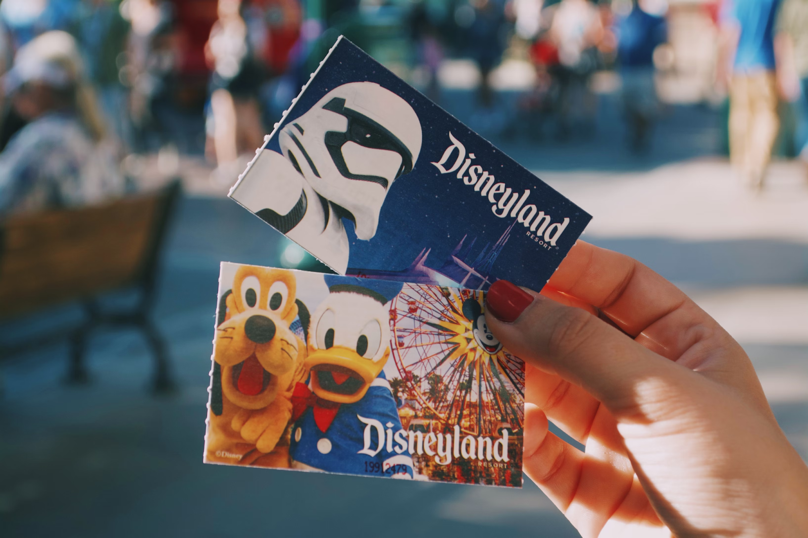 A child's hands holding two colorful Disneyland tickets, one featuring Pluto and the other Donald Duck, with the park's entrance blurred behind.
