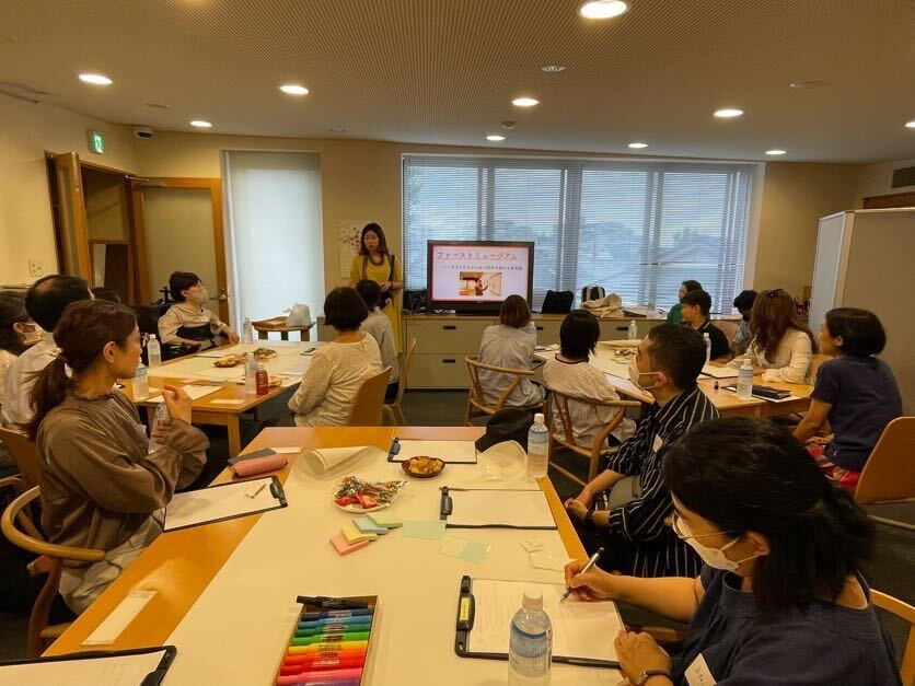 ちひろ美術館の３階のお部屋ぎゅうぎゅうになるくらいの人がいます。
３グループのテーブルを用意し、１グループあたり５〜６人ほどが座っています。中央には山田がモニターに写ったスライドを見せながらイントロダクションについて話している様子がわかります。参加者のみなさん真剣に聞いてくれている様子です。