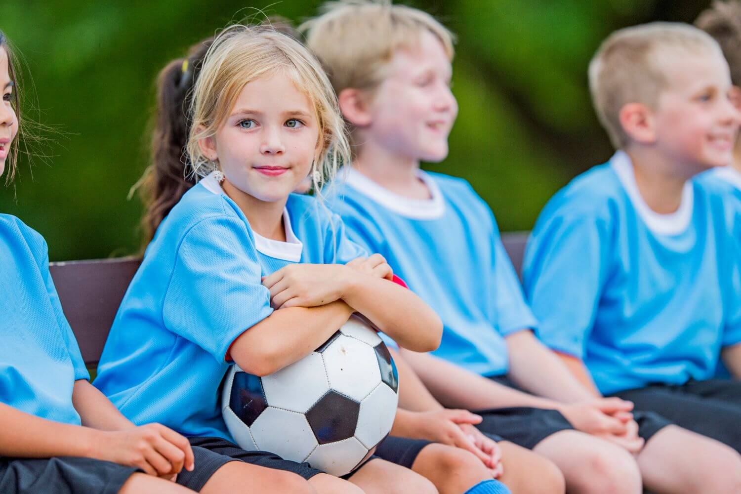 A team of youth coed soccer players in blue jerseys are sitting on a bench. A young girl is smiling while holding onto a soccer ball