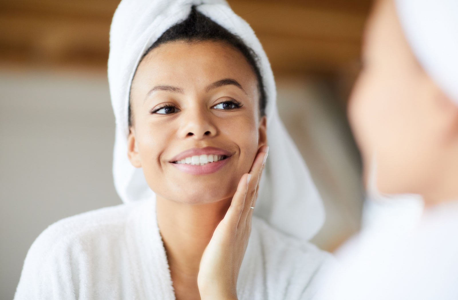A woman in a white bathrobe and a white towel wrapped around her head, looks at her clear skin in the mirror