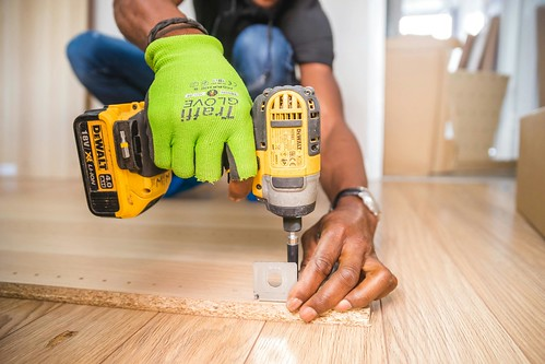 A man wearing a green glove on his right hand kneels over a wooden plank and drills a hole into it - What is a Trade School? Everything You Need to Know