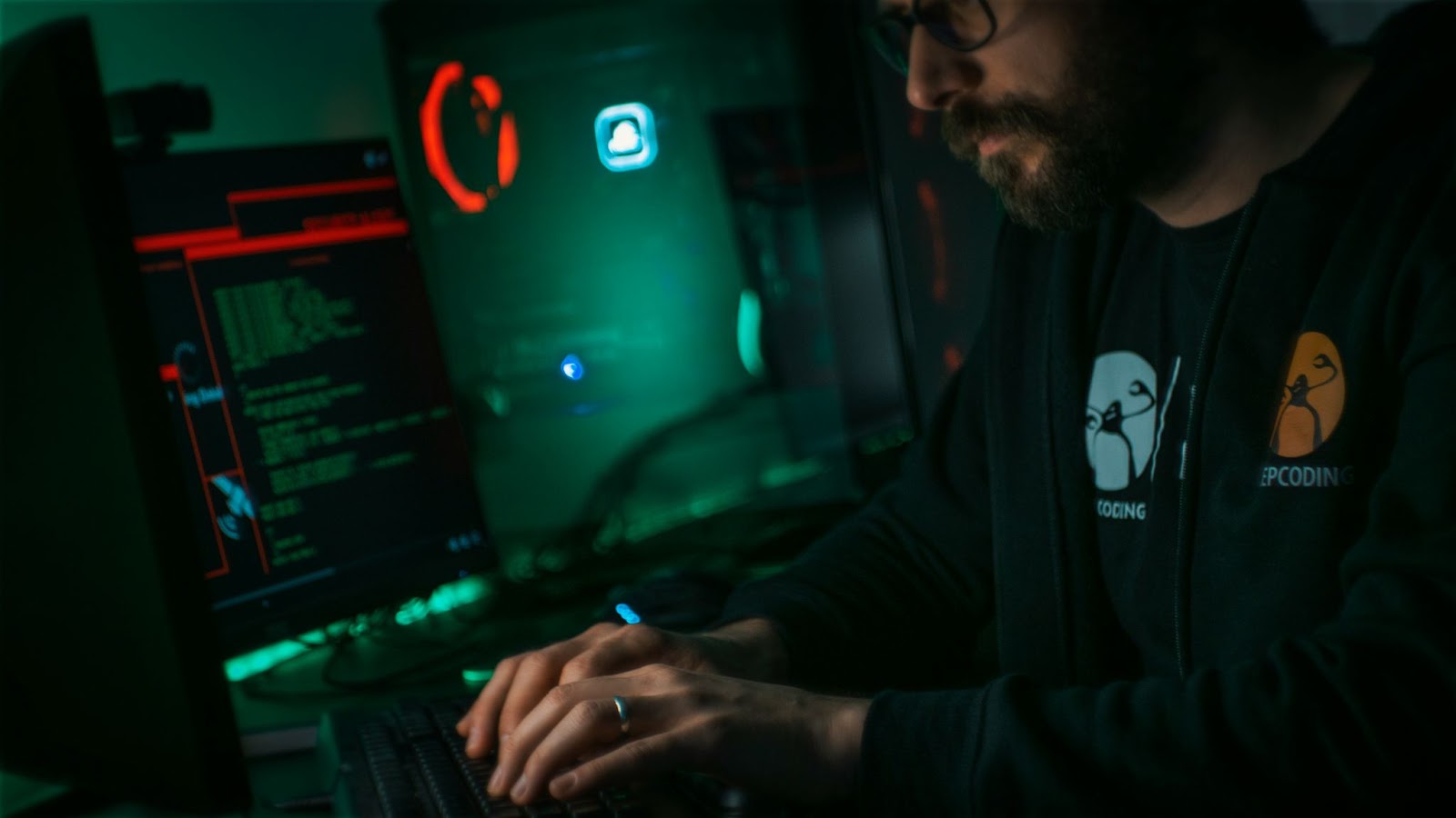 A man in a semi-dark room stands in front of his computer screens and writes on the keyboard