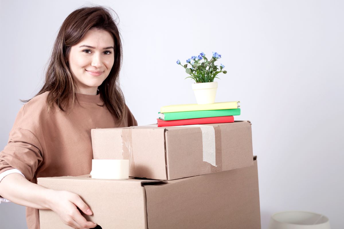 Student moving into a new space, holding cardboard boxes with notebooks and a plant.