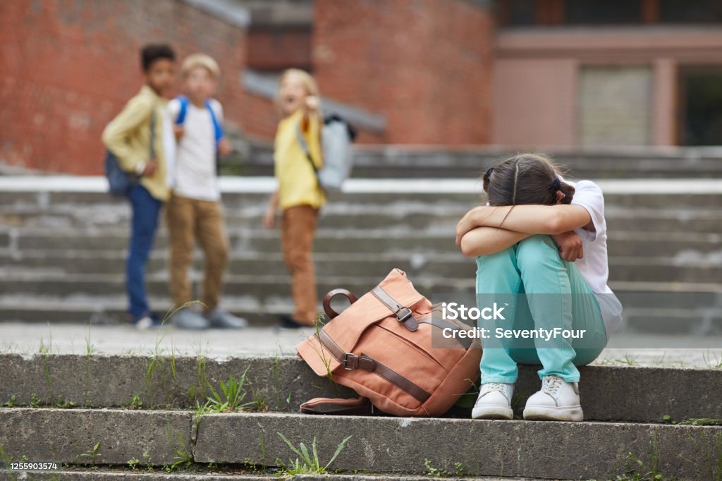 School Bullying Full length portrait of crying schoolgirl sitting on stairs outdoors with group of teasing children bullying her in background, copy space Bullying Stock Photo