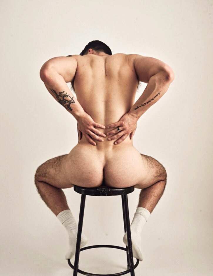 Dillon Cassidy sitting naked on a stool with his back to the camera showing off his muscled ass and tattoos