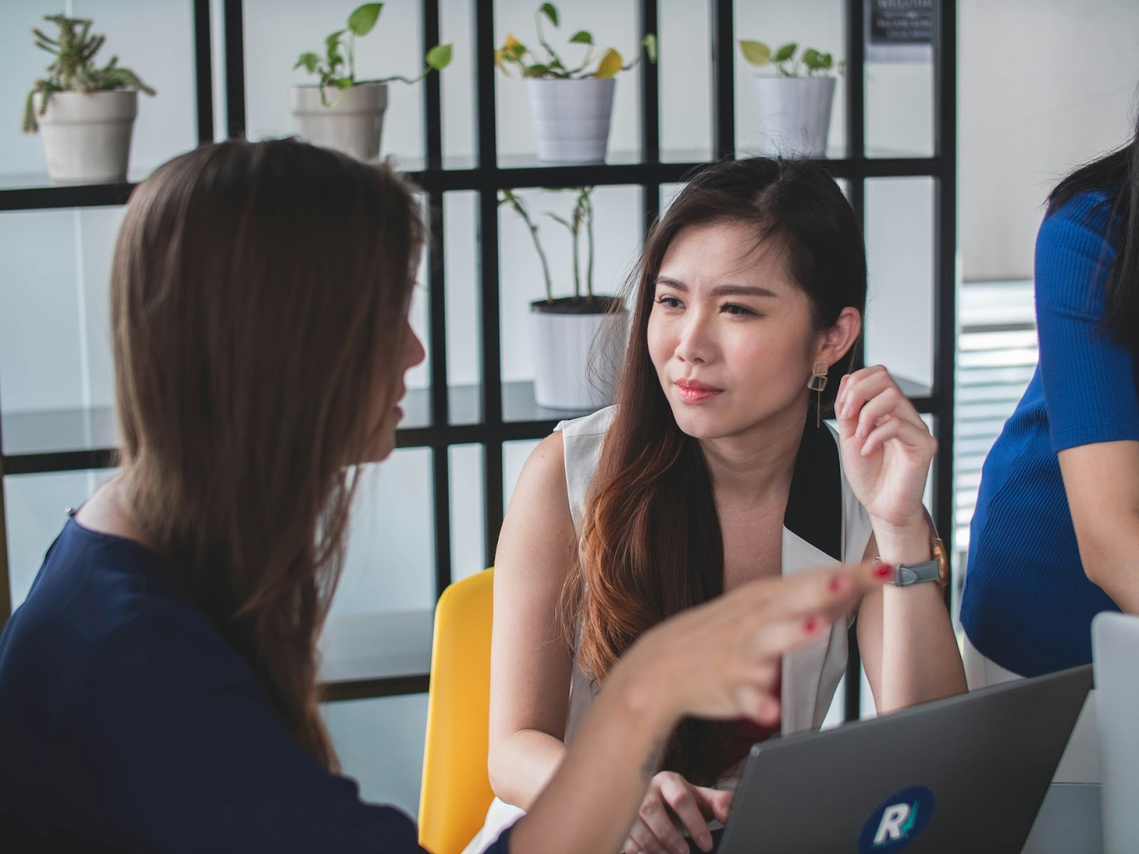 Two women discussing work in an office, one holding a pen and the other looking at a laptop screen.