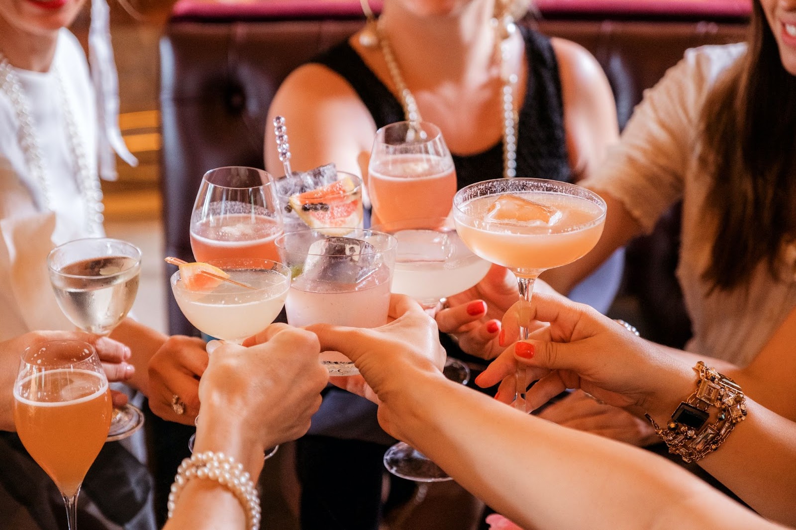 A group of women cheerfully clinking glasses filled with various pink and orange cocktails, symbolizing friendship and celebration.