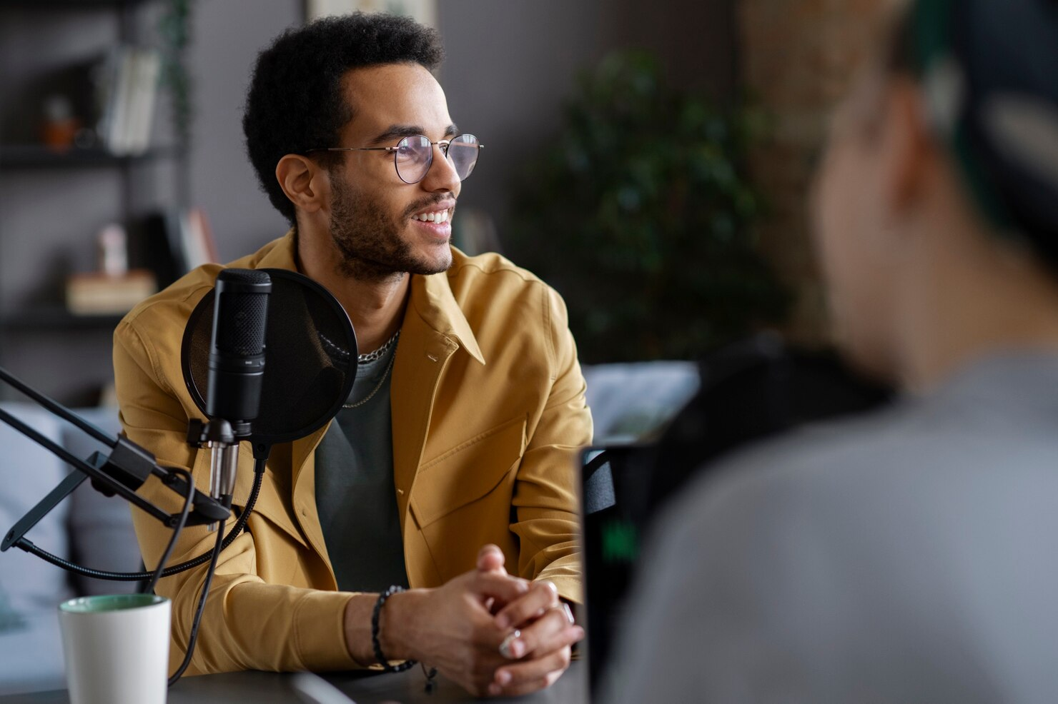 homem jovem negro, conversa com outro homem desfocado na frente de um microfone profissional durante a gravação de um podcast independente. Ele usa óculos, uma camisa cinza e por cima um blazer marrom claro