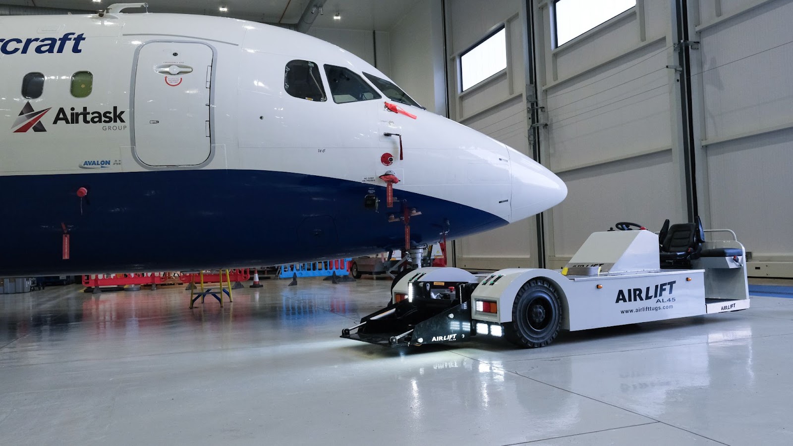 A white electric towing vehicle parked in front of a large blue and white research aircraft inside a hangar. The white lights on the tug are illuminating the hangar floor around it.