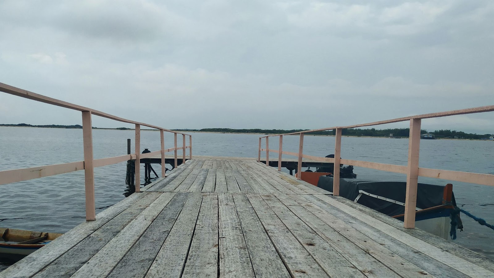 Pier de madeira centralizado na imagem, com a lagoa em segundo plano. Dia nublado e cinza.