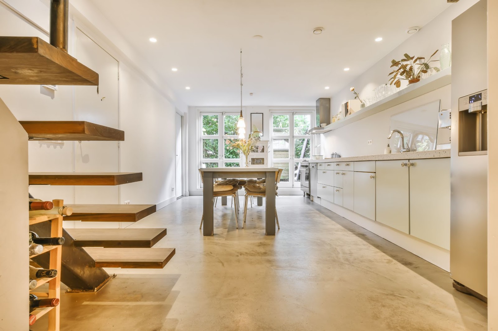 : A sleek and stylish dining area in cream and light brown hues featuring stained concrete floors, a wooden dining table in the center, shelves, and wooden stairs on the side, with glass doors in the background, creating a spacious feel.