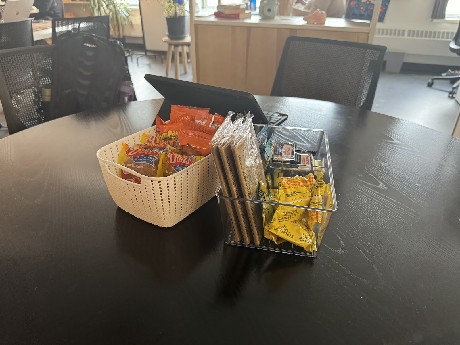 A white basket with cookies and Bear Paws is next to a clear container with popcorn and other snacks. They are on a black wood table.