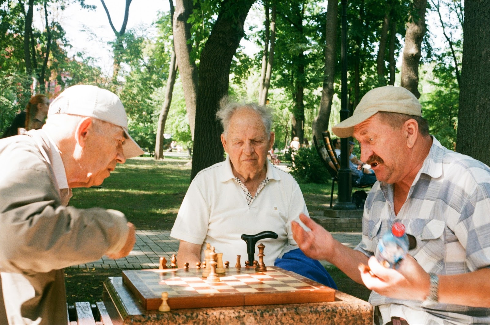  Three men sitting at a park. Two are playing chess and one man is watching