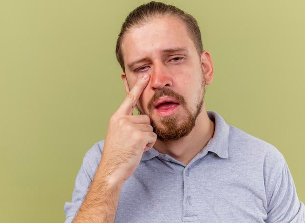 Close-up de um jovem bonito doente e fraco colocando o dedo embaixo do olho, olhando para a frente, isolado na parede verde oliva