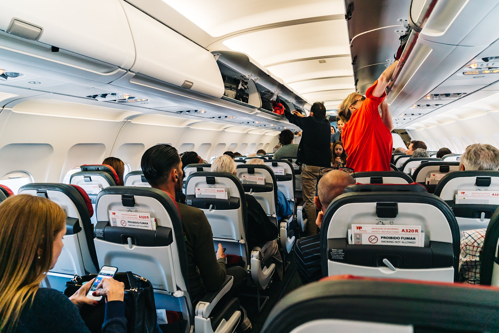 An inside the cabin view of economy class of TAP Air Portugal that shows a two seated rows with a cramped over the head luggage storage area.