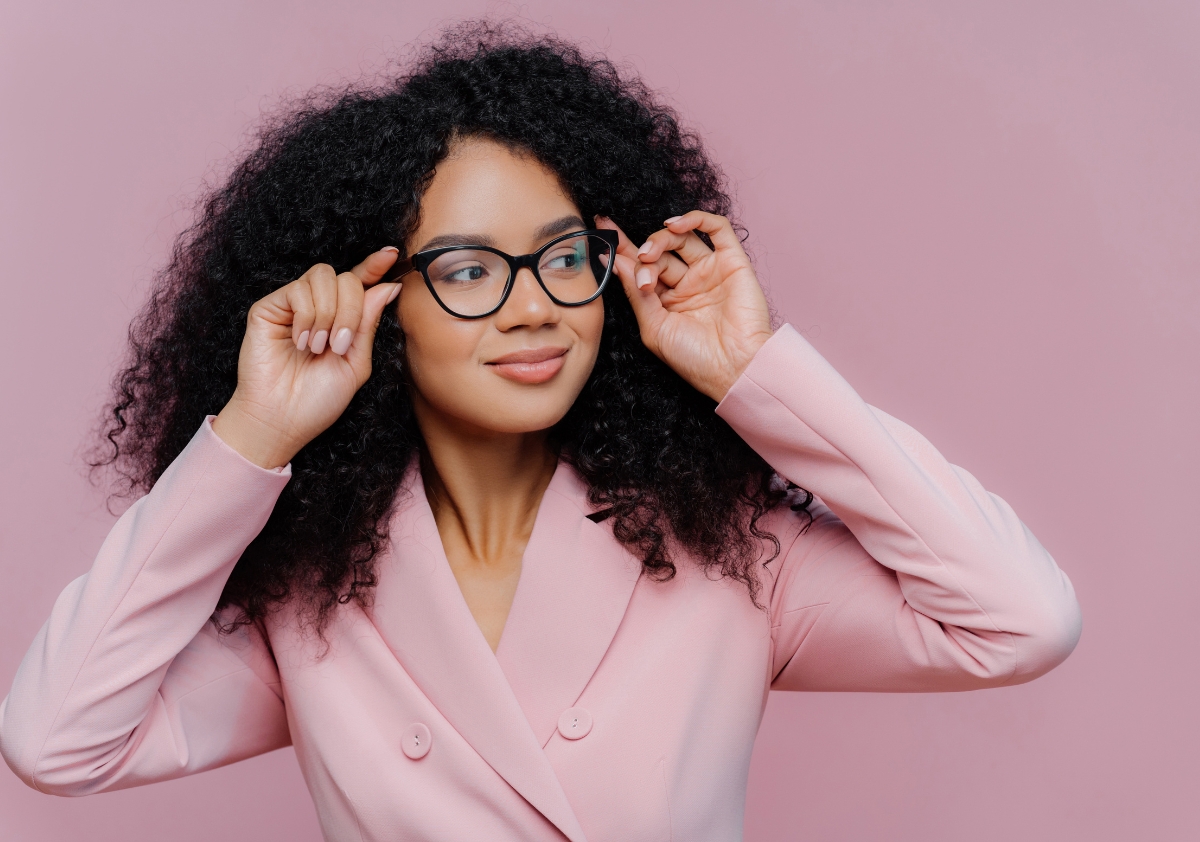 Woman of color with curly hair wearing a baby pink suit and glasses standing in front of a baby pink backdrop posing for Women's History Month.