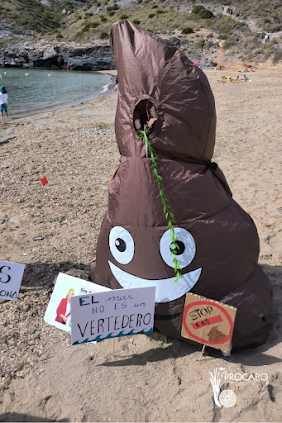 Imagen de una caca sonriente de whatsapp hecha con tela marrón en una playa (un cono marrón),  A sus pies hay pancatas clavadas en la arena que dicen "El mar no es un vertedero", "stop kks"