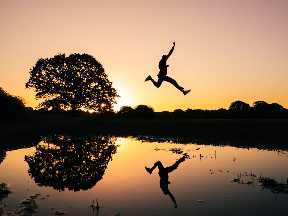 A Jumping Man and His Reflection in a Water Body With a Tree in the Background