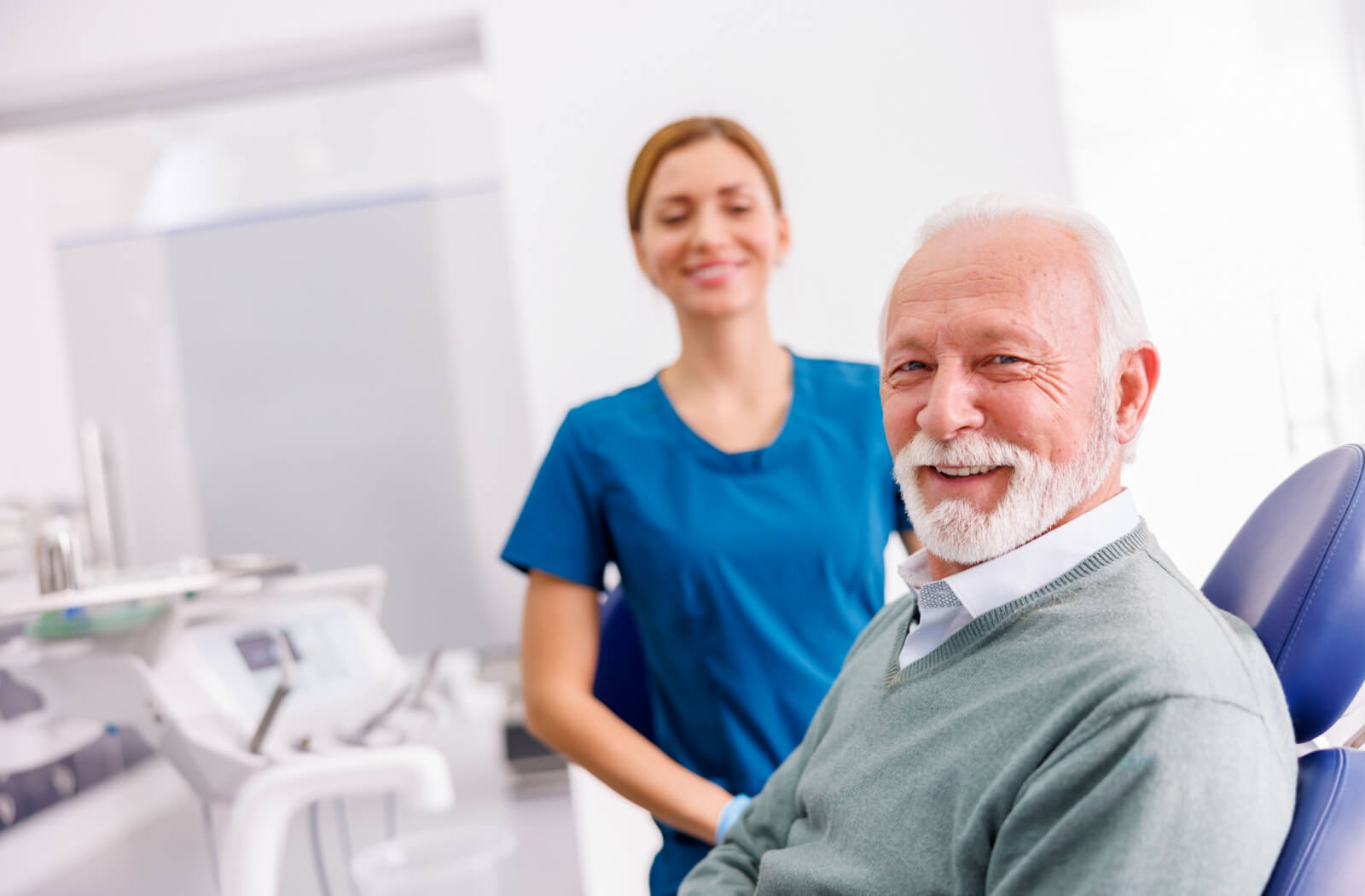 A senior man smiling and looking directly at the camera at a dental office with his dentist smiling in the background.
