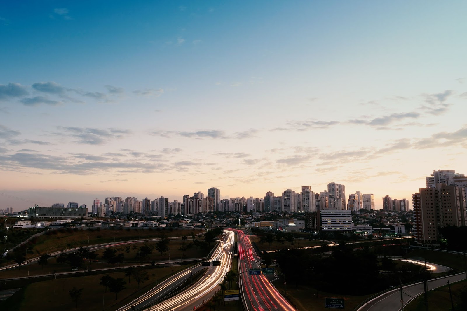 Vista panorâmica da cidade de São José dos Campos. Rodovia dividida pelo fluxo de ida e vinda, com as luzes dos veículos formando um borrão. Céu azul com poucas nuvens.