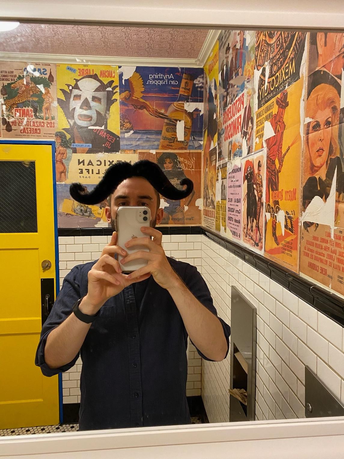 Stephan poses for a bathroom mirror selfie. A faux mustache stuck to the mirror is positioned so it looks like a floppy hairstyle on top of Stephan's head.