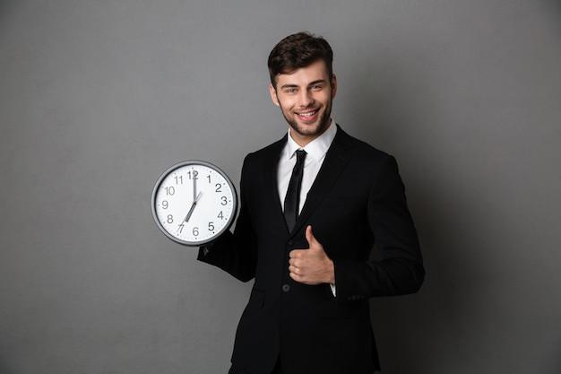 Young successful business man holding clock while showing thumb up gesture and 