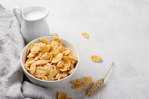 A bowl of cornflakes cereal with a glass of milk on the side