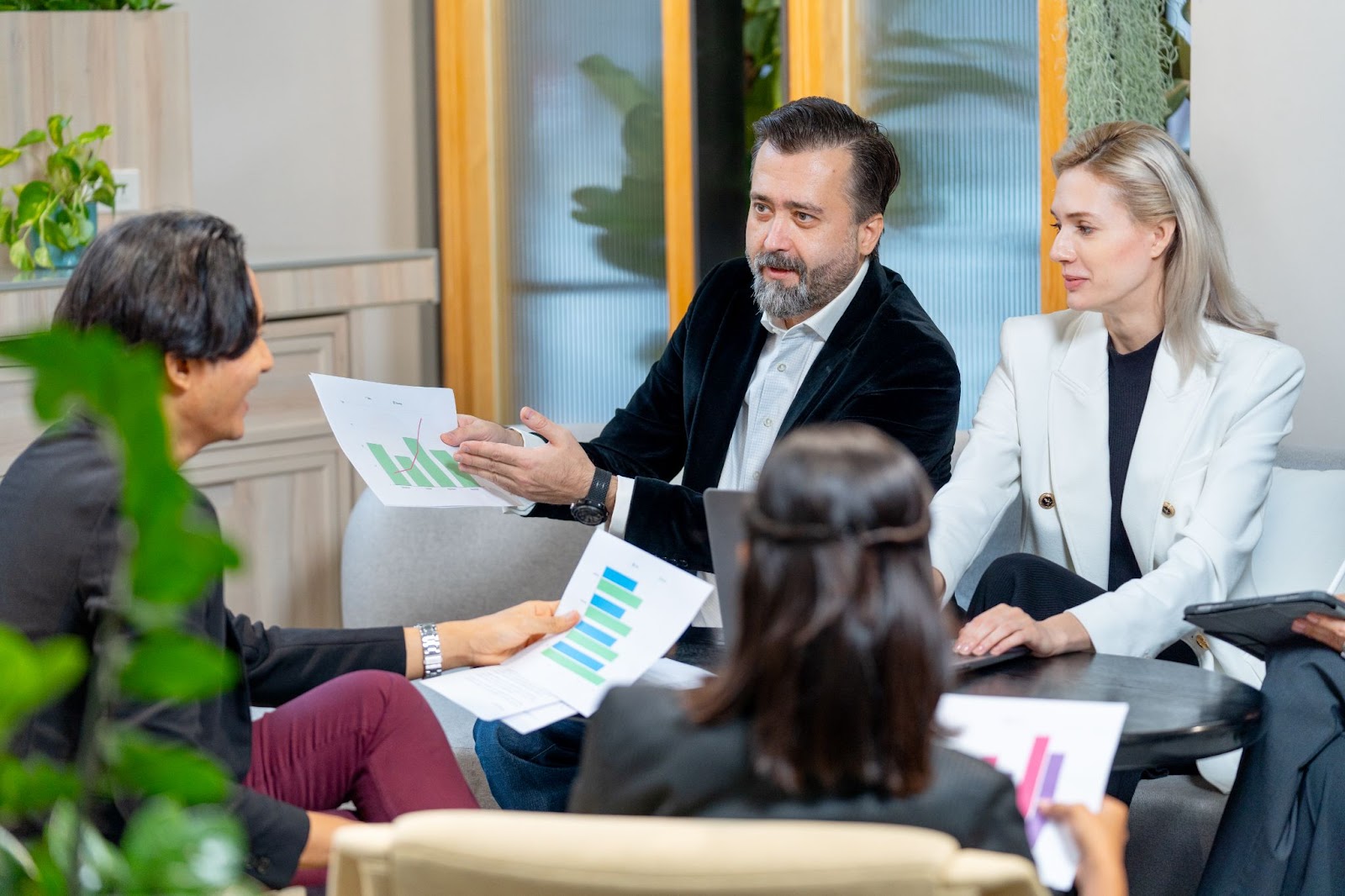 An Asian businessman discusses with a Caucasian male manager from a management consulting firm while two female executives listen intently. Lush greenery adorns the space, with a stylish glass door framed in wood in the background.