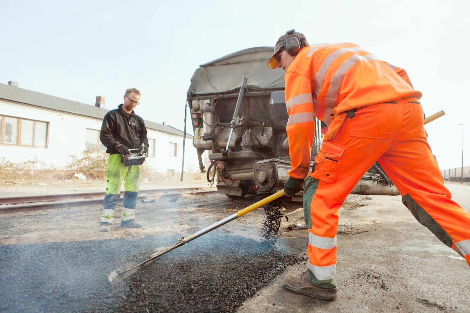 A manual worker wearing neon orange protective gear paves a road at a construction site while another worker looks on with a leveler.