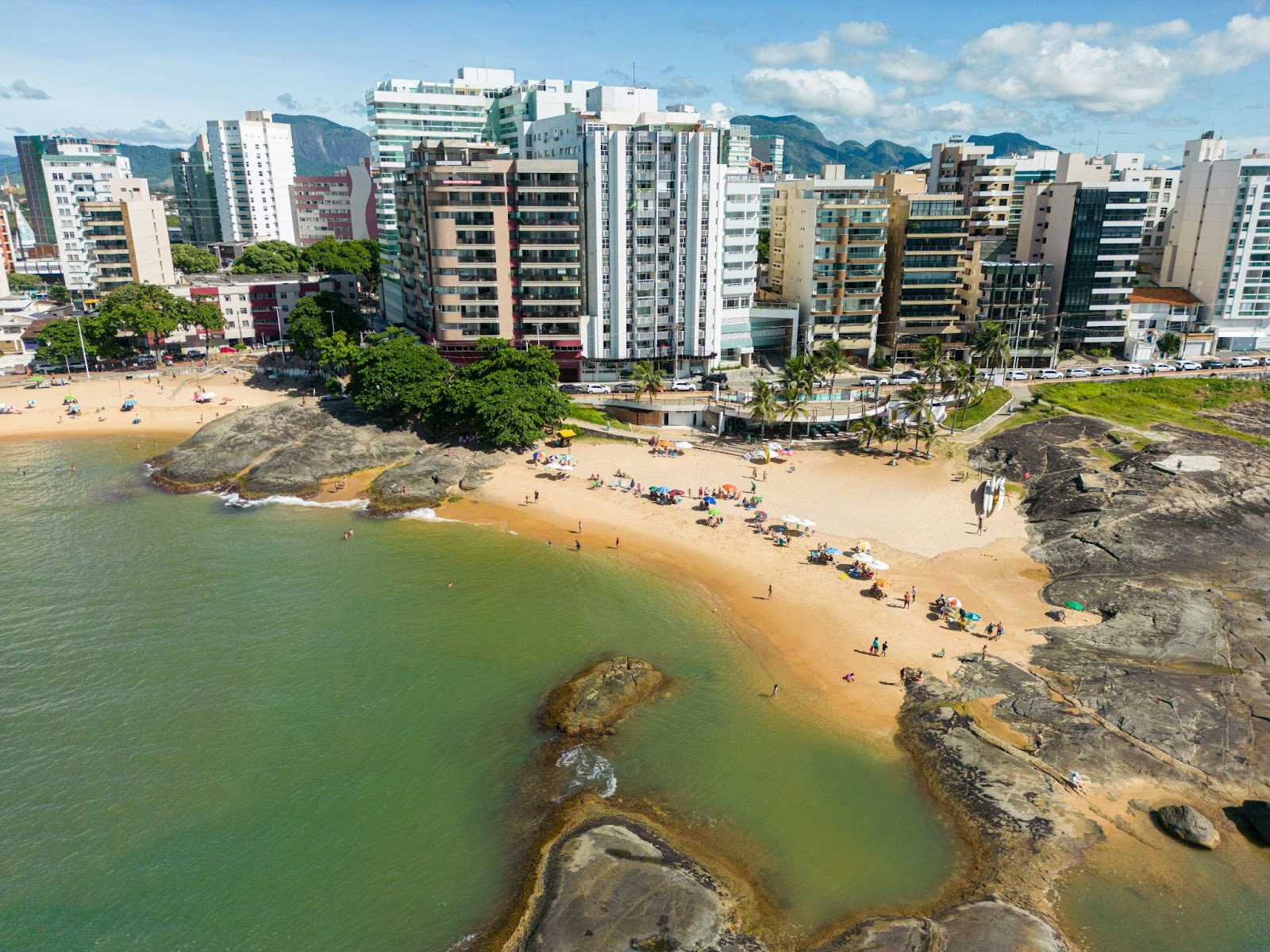 Vista aérea da Praia dos Namorados. Pequena faixa de areia entre rochas, com prédios da cidade de diferentes tamanhos atrás.