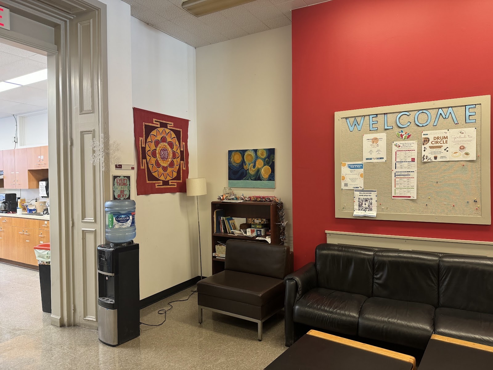 A room in the Multi-faith and Spirituality Centre office. There is a black leather couch, a bulletin board with some flyers and “Welcome” written in blue letters. There is a small bookcase, a water cooler and a bit of the kitchen is visible in the background.