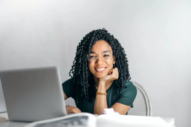 A young woman with glasses sitting at her desk, happily working on her laptop while making eye contact with the camera - How to Start a Cover Letter for Teens