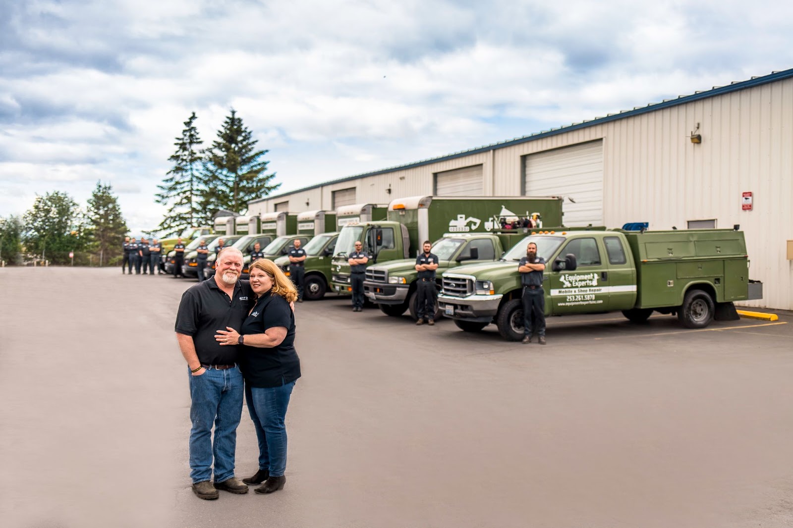 Equipment Experts, Inc. founders embracing in the foreground with trucks and mechanics lined up next to trucks in the background