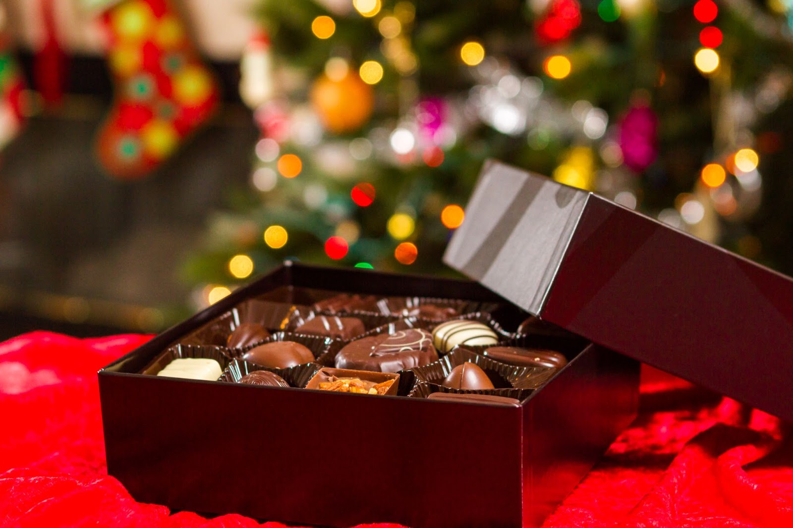 An opened box of chocolates, presented as a Christmas gift, with a softly blurred background of a twinkling Christmas tree and stockings
