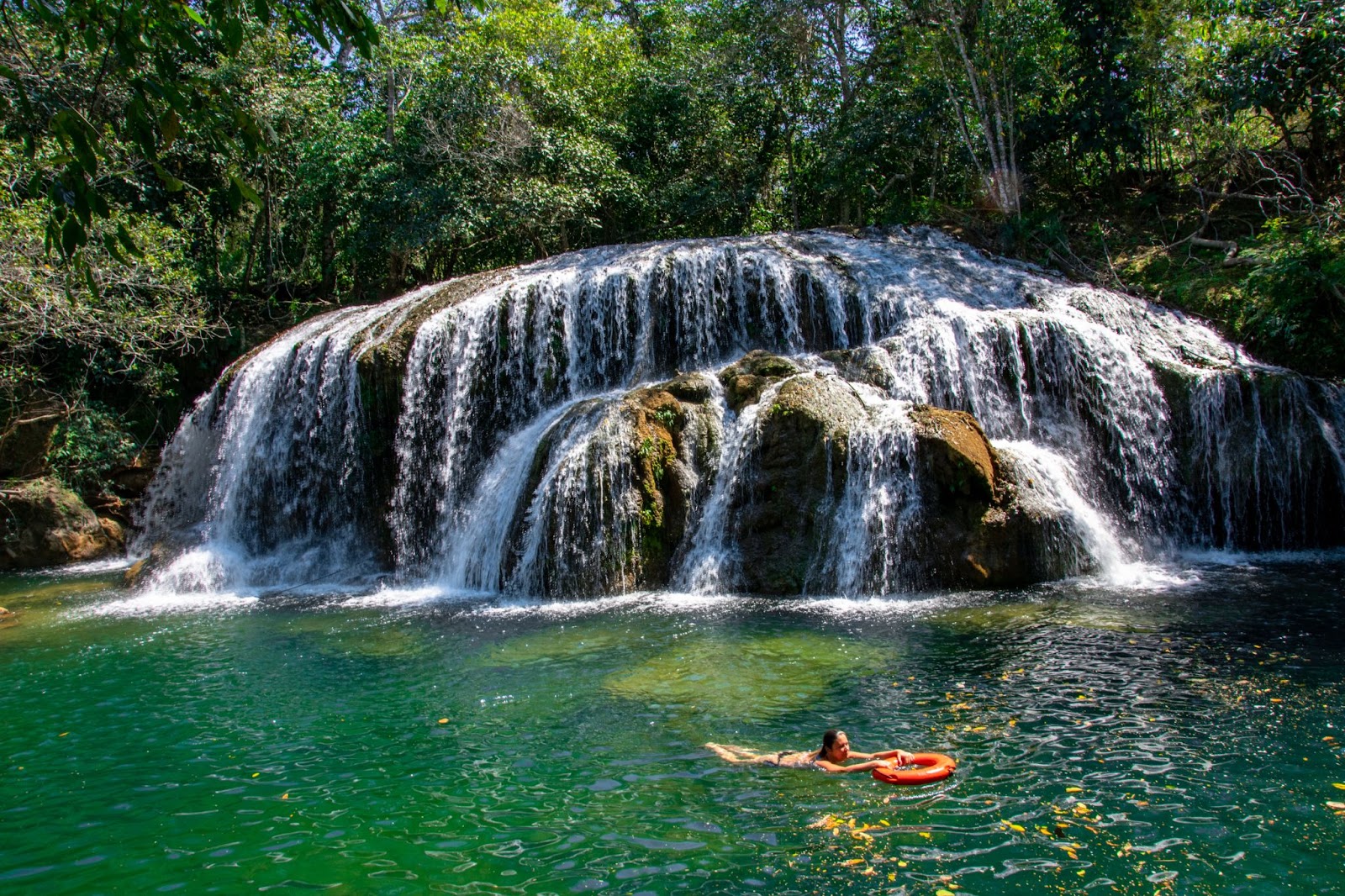 Turista nadando em uma cachoeira de Bonito, MS. A água desliza por grandes pedras marrons e desce até um poço esverdeado. Ao redor da cascata, aparecem trechos de floresta verde e densa