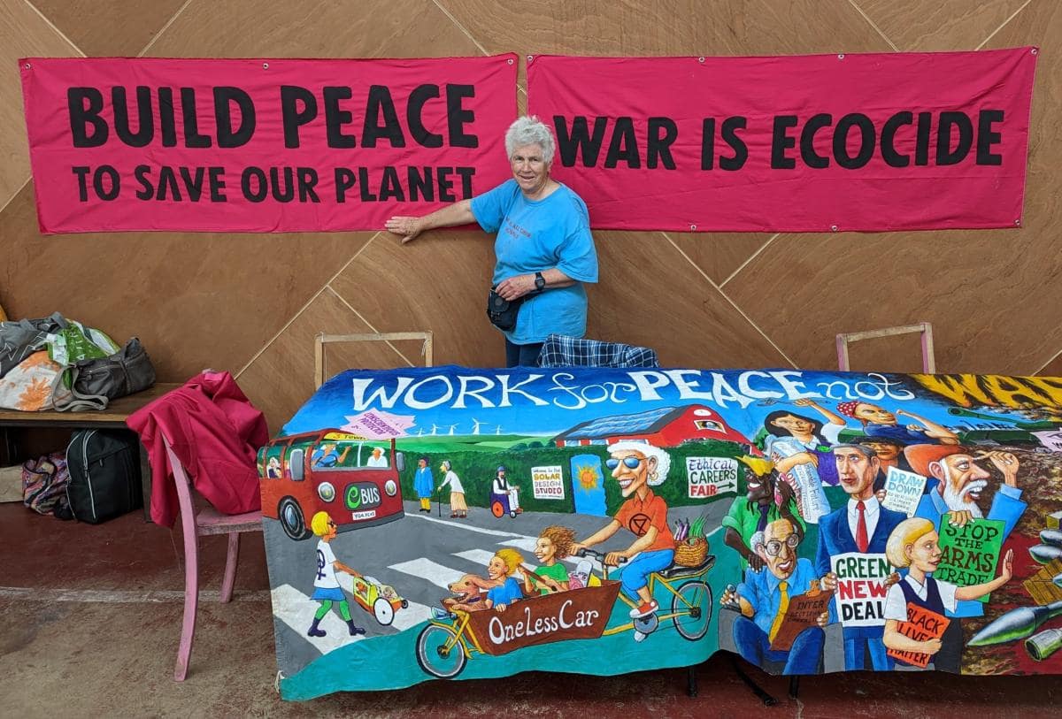 A rebel stands among banners, one a beautiful artwork showing an eco-village, the others talking of peace saving our planet, and war being ecocide