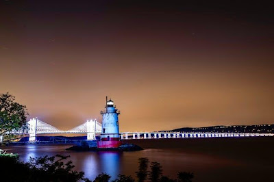 Nighttime scene with a lighthouse and illuminated bridge over tranquil waters in Westchester, NY.