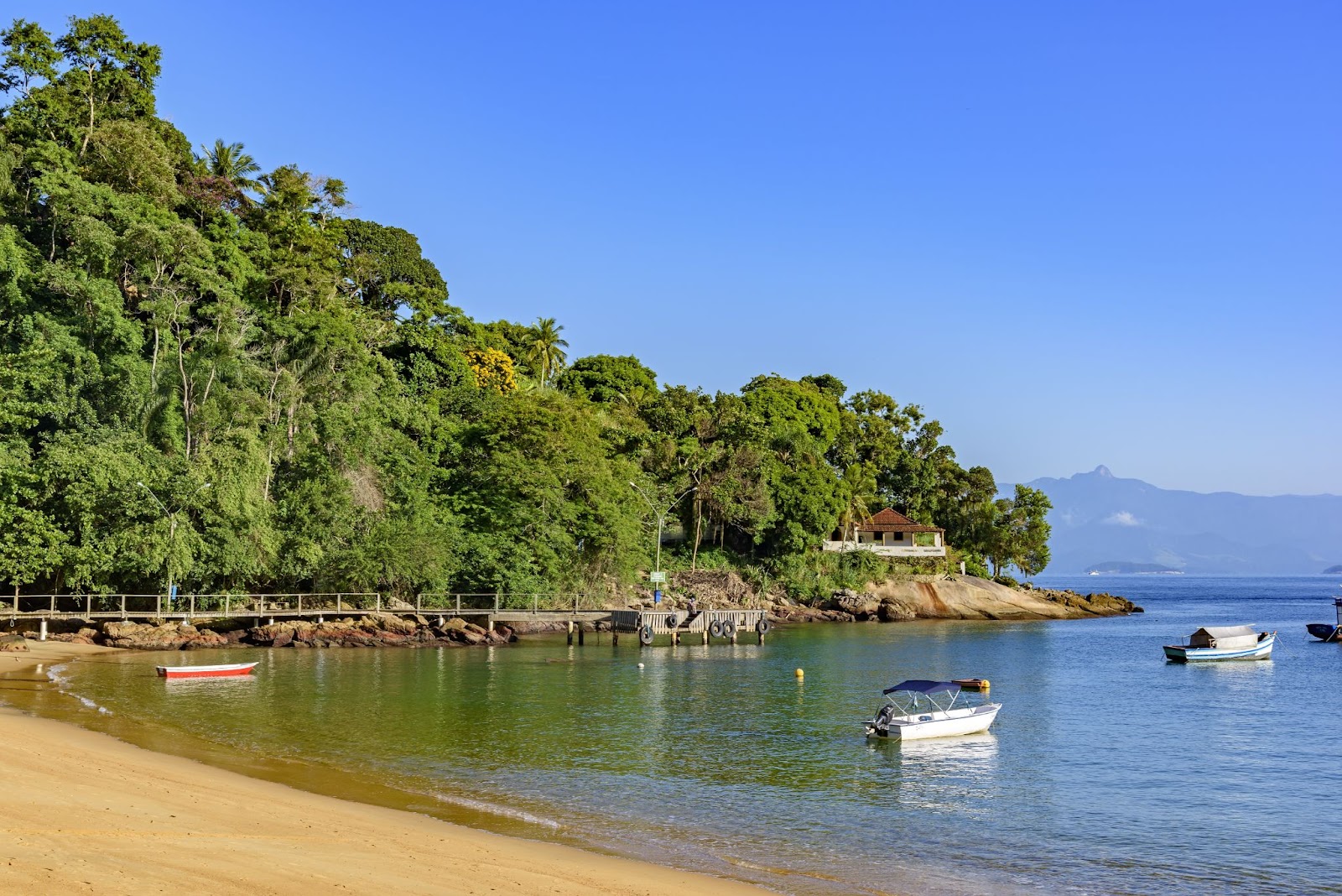 Final da Praia Vermelha da Ilha Grande, em Ilha Grande, Rio de Janeiro. A faixa de areia dourada termina em uma ponta coberta por floresta nativa. Alguns barquinhos flutuam na água de tonalidade verde profunda