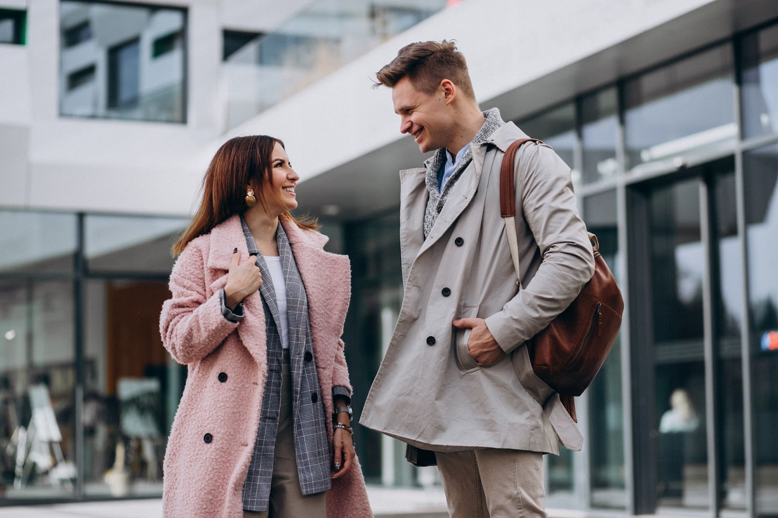 Man and woman standing outside a building wearing winter coats and sharing a joke.