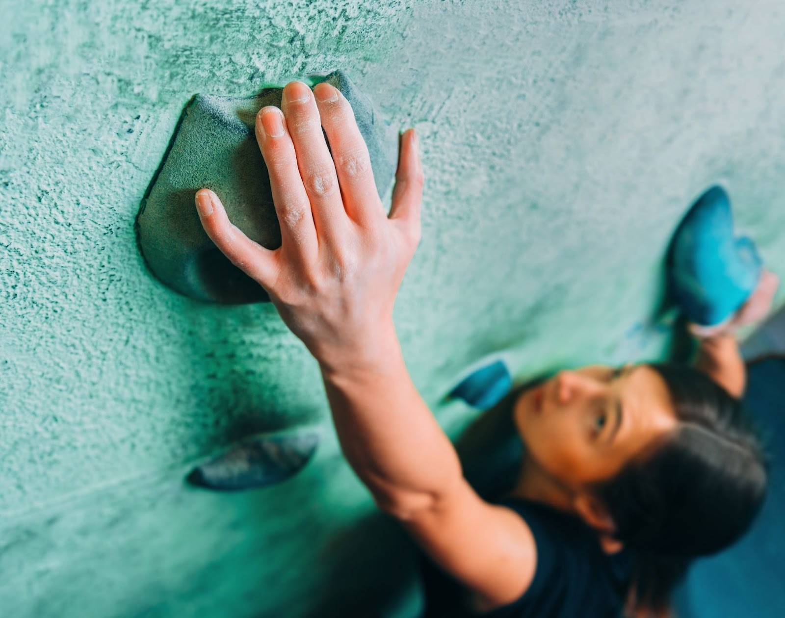 Mulher em um paredão de escalada, com sua mão direita em foco e o restante de seu corpo desfocado.